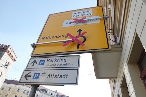 A set of road signs are mounted on a pole next to a building. The top sign is large and yellow with arrows and text that have been crossed out with red tape, indicating direction changes. Below it, there are two smaller blue and white signs pointing towards parking areas and an old town center. An older brick building and other classical structures are partly visible in the background against a clear sky.
