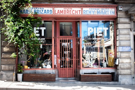 A storefront with large glass windows displaying the name 'PIET' and the website 'moodshop.com'. The facade features vintage signage with names like 'Huis Lambrecht', 'Marie Brizard', and 'Remy Martin'. Ivy grows on the left side, and there is a wooden bench below the windows. The building appears to be part of an urban street scene with some reflections in the glass.