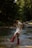 A person wearing a white dress and red boots is playfully splashing water in a forest stream. Surrounding them are lush, green trees, and the sunlight filters through the canopy, casting dappled shadows on the water.