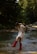 A person wearing a white dress and red boots is playfully splashing water in a forest stream. Surrounding them are lush, green trees, and the sunlight filters through the canopy, casting dappled shadows on the water.