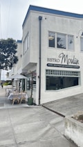A corner building with a sign for Bistro Moulin, specializing in wine, boutique, and European cuisine. Outside, there are tables set with white tablecloths under a white umbrella, surrounded by some planters with flowers. The street is quiet and overcast, with a paved sidewalk and some greenery nearby.