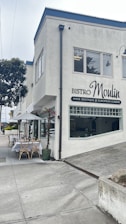 A corner building with a sign for Bistro Moulin, specializing in wine, boutique, and European cuisine. Outside, there are tables set with white tablecloths under a white umbrella, surrounded by some planters with flowers. The street is quiet and overcast, with a paved sidewalk and some greenery nearby.