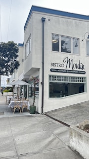 A corner building with a sign for Bistro Moulin, specializing in wine, boutique, and European cuisine. Outside, there are tables set with white tablecloths under a white umbrella, surrounded by some planters with flowers. The street is quiet and overcast, with a paved sidewalk and some greenery nearby.
