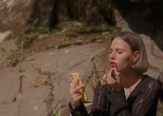 A person is sitting outdoors on a rocky surface, applying lipstick. They are using a yellow phone as a mirror and wearing a black lace top. The background includes stone and some small green plants.