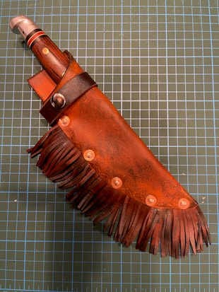 A knife with a brown leather sheath featuring fringe and rivet details. The handle is wooden with metallic accents, and the sheath has a dark strap and a stud for securing the blade. The background is a grid cutting mat.