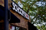 A building facade with a prominent sign that reads 'SCHOOL OF ROCK' in bold black and white letters on a brick wall. The logo incorporates a red symbol where the 'O' would be, which is stylized with a musical note. Lush green trees partially shade the building, creating a pleasant outdoor atmosphere.