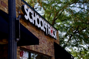 A building facade with a prominent sign that reads 'SCHOOL OF ROCK' in bold black and white letters on a brick wall. The logo incorporates a red symbol where the 'O' would be, which is stylized with a musical note. Lush green trees partially shade the building, creating a pleasant outdoor atmosphere.