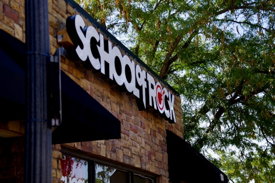 A building facade with a prominent sign that reads 'SCHOOL OF ROCK' in bold black and white letters on a brick wall. The logo incorporates a red symbol where the 'O' would be, which is stylized with a musical note. Lush green trees partially shade the building, creating a pleasant outdoor atmosphere.