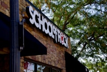 A building facade with a prominent sign that reads 'SCHOOL OF ROCK' in bold black and white letters on a brick wall. The logo incorporates a red symbol where the 'O' would be, which is stylized with a musical note. Lush green trees partially shade the building, creating a pleasant outdoor atmosphere.
