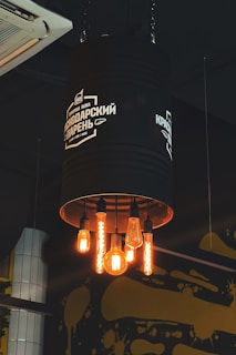 A creative industrial-style light fixture fashioned from a large, black metal cylinder hanging from the ceiling, with five exposed, vintage-style light bulbs glowing warmly. The fixture bears white text and a logo, and there is an air conditioning unit and artistic yellow paint splatters on the wall in the background.