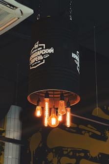 A creative industrial-style light fixture fashioned from a large, black metal cylinder hanging from the ceiling, with five exposed, vintage-style light bulbs glowing warmly. The fixture bears white text and a logo, and there is an air conditioning unit and artistic yellow paint splatters on the wall in the background.