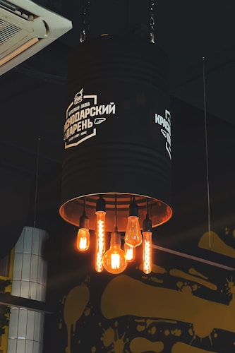 A creative industrial-style light fixture fashioned from a large, black metal cylinder hanging from the ceiling, with five exposed, vintage-style light bulbs glowing warmly. The fixture bears white text and a logo, and there is an air conditioning unit and artistic yellow paint splatters on the wall in the background.