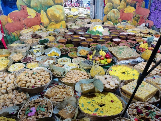 A large assortment of traditional Indian sweets and savories arranged on a table. There are numerous bowls and plates filled with various kinds of snacks, each crafted with care and arranged in an inviting manner. The backdrop includes colorful paintings of cattle against a backdrop of rocks and flowers.