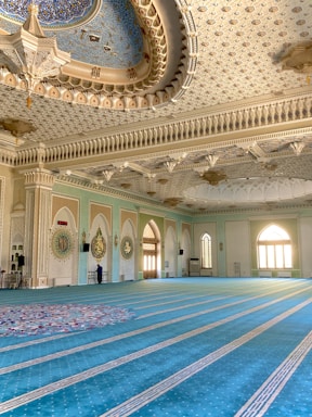 An intricately decorated mosque interior featuring a grand chandelier and beautifully patterned ceiling. The spacious room has a blue patterned carpet and walls adorned with Arabic calligraphy and decorative motifs. Natural light streams in through arched windows, creating a serene atmosphere.
