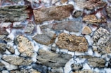 A textured wall made from variously sized and shaped natural stones held together with visible mortar. The stones exhibit diverse earth tones including brown, gray, and beige, and some stones have a distinct rough surface. The mortar shows slight discoloration, with hints of white and red mixed in.