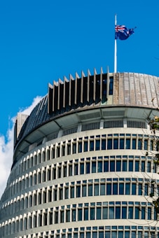 A modern cylindrical building with a unique architectural design featuring multiple horizontal rows of windows. The top of the structure has a serrated edge and a flagpole with a New Zealand flag fluttering against a clear blue sky.
