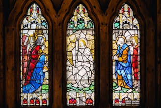 Three arched stained glass windows depict biblical figures in vibrant colors. The central panel features an angel with wings, surrounded by kneeling figures in blue and red robes, each with a halo. Ornate patterns and intricate architectural designs enhance the background.