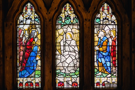 Three arched stained glass windows depict biblical figures in vibrant colors. The central panel features an angel with wings, surrounded by kneeling figures in blue and red robes, each with a halo. Ornate patterns and intricate architectural designs enhance the background.