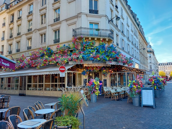 A charming street corner caf&eacute; with a vibrant floral display covering the entrance and extending along the building. Tables and chairs are set outside on the cobblestone pavement, inviting patrons to sit. The surroundings feature classic European architecture with tall windows and balconies.