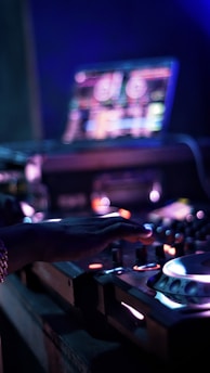 A DJ is operating a mixing console with one hand, surrounded by colorful lights. The background features a blurred laptop screen displaying music software.