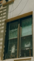 A double-pane window with a brown frame is installed in a light-colored wall. The panes are slightly dirty and show reflections and diagonal patterns. Behind the window, white fabric drapes hang in folds. An electrical wire runs across the top corner of the wall, casting a shadow.