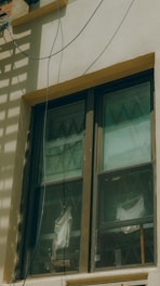 A double-pane window with a brown frame is installed in a light-colored wall. The panes are slightly dirty and show reflections and diagonal patterns. Behind the window, white fabric drapes hang in folds. An electrical wire runs across the top corner of the wall, casting a shadow.