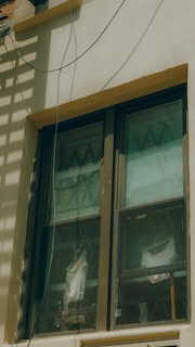 A double-pane window with a brown frame is installed in a light-colored wall. The panes are slightly dirty and show reflections and diagonal patterns. Behind the window, white fabric drapes hang in folds. An electrical wire runs across the top corner of the wall, casting a shadow.