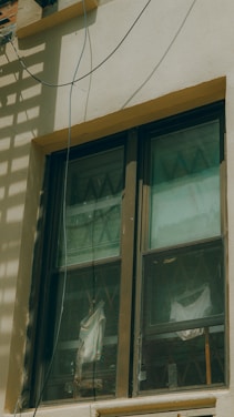 A double-pane window with a brown frame is installed in a light-colored wall. The panes are slightly dirty and show reflections and diagonal patterns. Behind the window, white fabric drapes hang in folds. An electrical wire runs across the top corner of the wall, casting a shadow.