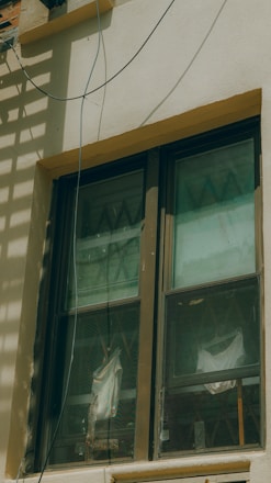 A double-pane window with a brown frame is installed in a light-colored wall. The panes are slightly dirty and show reflections and diagonal patterns. Behind the window, white fabric drapes hang in folds. An electrical wire runs across the top corner of the wall, casting a shadow.
