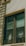 A double-pane window with a brown frame is installed in a light-colored wall. The panes are slightly dirty and show reflections and diagonal patterns. Behind the window, white fabric drapes hang in folds. An electrical wire runs across the top corner of the wall, casting a shadow.