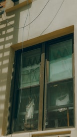 A double-pane window with a brown frame is installed in a light-colored wall. The panes are slightly dirty and show reflections and diagonal patterns. Behind the window, white fabric drapes hang in folds. An electrical wire runs across the top corner of the wall, casting a shadow.