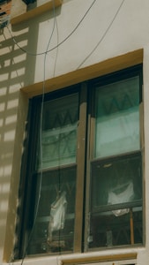 A double-pane window with a brown frame is installed in a light-colored wall. The panes are slightly dirty and show reflections and diagonal patterns. Behind the window, white fabric drapes hang in folds. An electrical wire runs across the top corner of the wall, casting a shadow.
