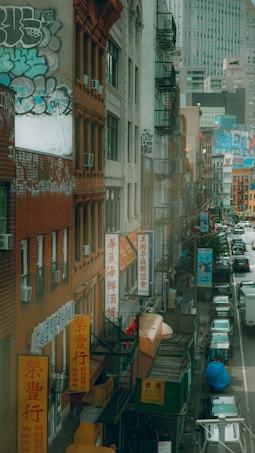A bustling urban street scene features tall buildings adorned with colorful graffiti and vibrant Chinese signage. Fire escapes line the side of the structures, and numerous vehicles and pedestrians fill the busy street below. The atmosphere is lively and reflective of a multicultural city area.