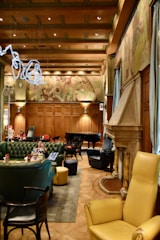 An elegant room with high wood-paneled ceilings and decorative wall art. The space features plush green and yellow leather seating, a grand piano, and a large stone fireplace. A woman is seated on a green sofa, and other patrons are visible in the background. The ambient lighting creates a cozy atmosphere.