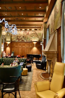 An elegant room with high wood-paneled ceilings and decorative wall art. The space features plush green and yellow leather seating, a grand piano, and a large stone fireplace. A woman is seated on a green sofa, and other patrons are visible in the background. The ambient lighting creates a cozy atmosphere.