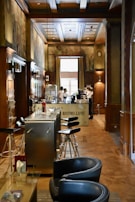 A cozy interior of a bistro with wooden paneling and decorative murals on the walls. Modern bar stools line a sleek metal counter, and there are two servers attending to the area near a coffee machine. Soft natural light filters in through a large window, enhancing the warm and inviting atmosphere.