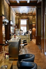 A cozy interior of a bistro with wooden paneling and decorative murals on the walls. Modern bar stools line a sleek metal counter, and there are two servers attending to the area near a coffee machine. Soft natural light filters in through a large window, enhancing the warm and inviting atmosphere.