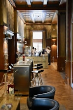 A cozy interior of a bistro with wooden paneling and decorative murals on the walls. Modern bar stools line a sleek metal counter, and there are two servers attending to the area near a coffee machine. Soft natural light filters in through a large window, enhancing the warm and inviting atmosphere.