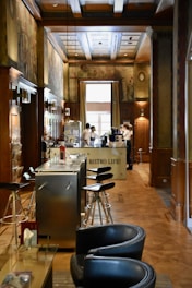A cozy interior of a bistro with wooden paneling and decorative murals on the walls. Modern bar stools line a sleek metal counter, and there are two servers attending to the area near a coffee machine. Soft natural light filters in through a large window, enhancing the warm and inviting atmosphere.