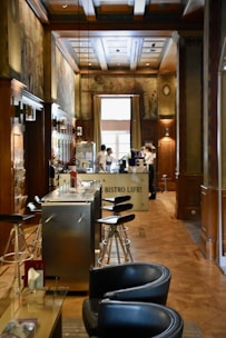 A cozy interior of a bistro with wooden paneling and decorative murals on the walls. Modern bar stools line a sleek metal counter, and there are two servers attending to the area near a coffee machine. Soft natural light filters in through a large window, enhancing the warm and inviting atmosphere.