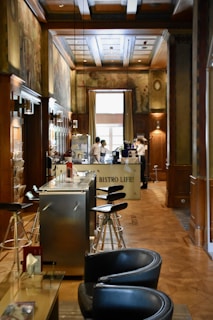 A cozy interior of a bistro with wooden paneling and decorative murals on the walls. Modern bar stools line a sleek metal counter, and there are two servers attending to the area near a coffee machine. Soft natural light filters in through a large window, enhancing the warm and inviting atmosphere.