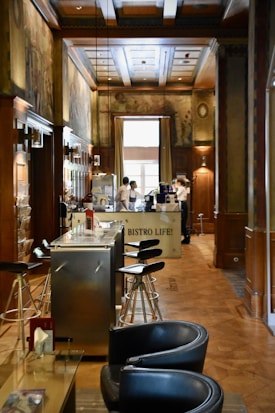 A cozy interior of a bistro with wooden paneling and decorative murals on the walls. Modern bar stools line a sleek metal counter, and there are two servers attending to the area near a coffee machine. Soft natural light filters in through a large window, enhancing the warm and inviting atmosphere.