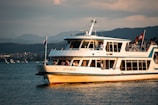 A white and blue passenger ferry named 'Uetliberg' is navigating on a body of water with people visible on board. The scene is set against a mountainous background, and the sky is partially covered with clouds. A Swiss flag is flying on the ferry.