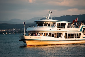 A white and blue passenger ferry named 'Uetliberg' is navigating on a body of water with people visible on board. The scene is set against a mountainous background, and the sky is partially covered with clouds. A Swiss flag is flying on the ferry.