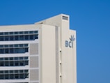 A modern building with multiple windows and a logo that includes the letters 'BCI' and a leaf motif. The structure is light-colored and set against a clear blue sky.