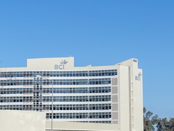 A large, modern office building with multiple floors and a predominantly glass facade. The building features the logo 'BCI' prominently on its exterior. The sky is a clear, bright blue, and there are some trees visible to the right of the structure.