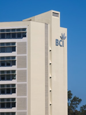 A tall beige building with rectangular windows and a large 'BCI' logo with a leaf symbol on the side. The building is set against a clear blue sky with some trees visible at the bottom right corner.
