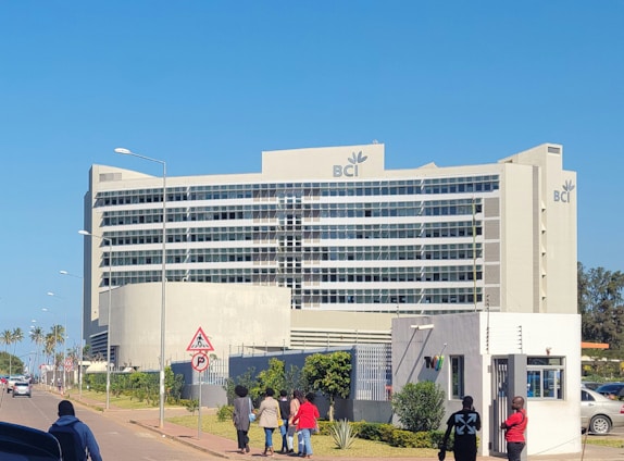 A modern, large, multi-story office building is prominently situated on a clear day, featuring the letters 'BCI' with a logo on its facade. The structure is surrounded by a well-maintained garden and a sidewalk where several people are walking. Nearby, cars are parked and some are driving along the street.
