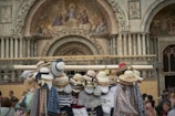A variety of stylish hats are displayed on a stand in front of an intricately decorated architectural structure. The building features ornate carvings and religious frescoes above a large archway. Several people are gathered nearby, looking at the items or walking past.