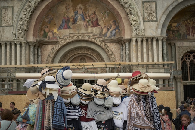 A variety of stylish hats are displayed on a stand in front of an intricately decorated architectural structure. The building features ornate carvings and religious frescoes above a large archway. Several people are gathered nearby, looking at the items or walking past.